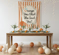 A styled party setup with a wooden table covered in orange-themed decorations, including oranges, greenery, and balloons. A large orange-and-white striped banner in the background reads 'Orange You Glad She Said Yes?' with a small decorative illustration.