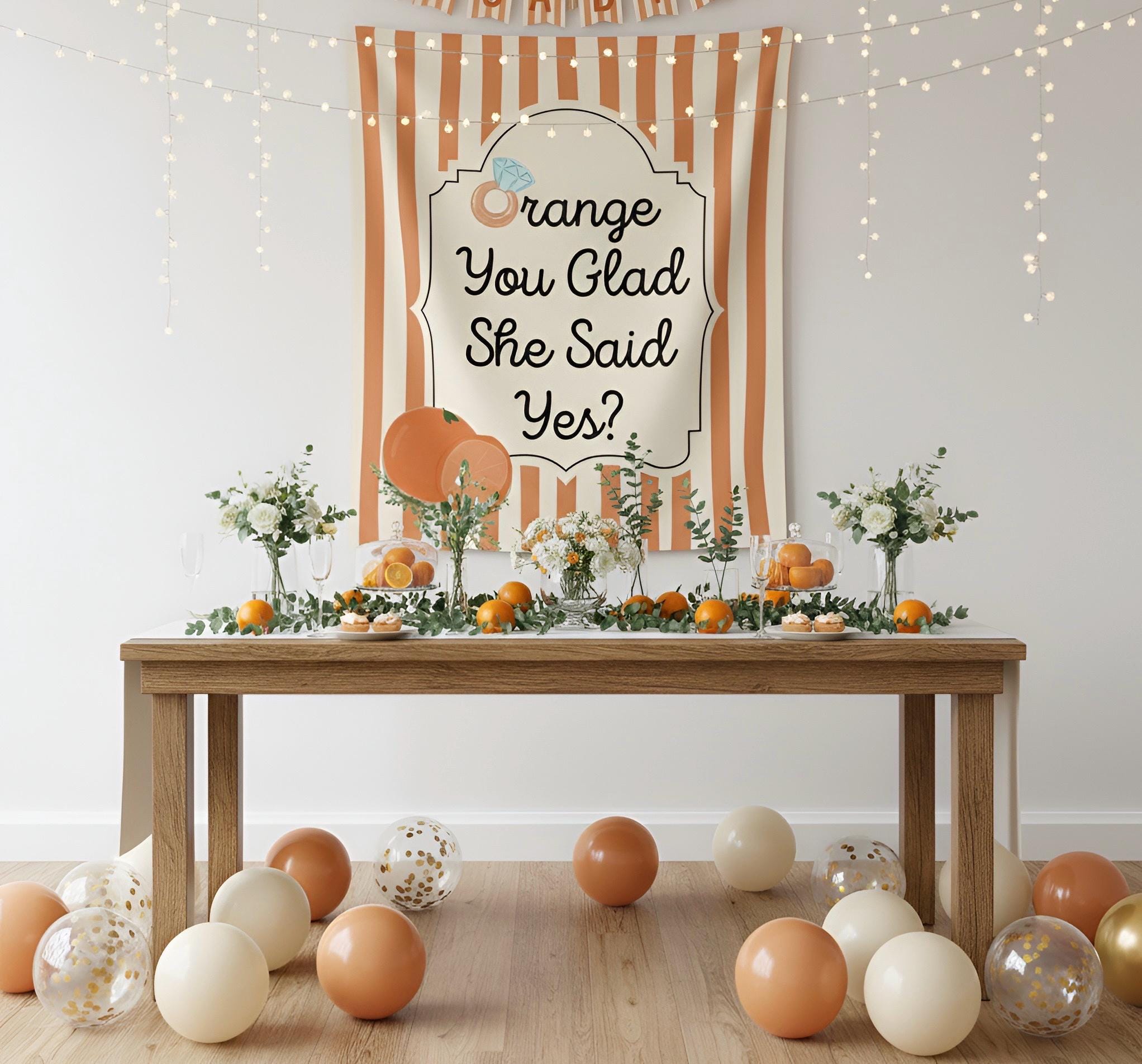 A styled party setup with a wooden table covered in orange-themed decorations, including oranges, greenery, and balloons. A large orange-and-white striped banner in the background reads 'Orange You Glad She Said Yes?' with a small decorative illustration.