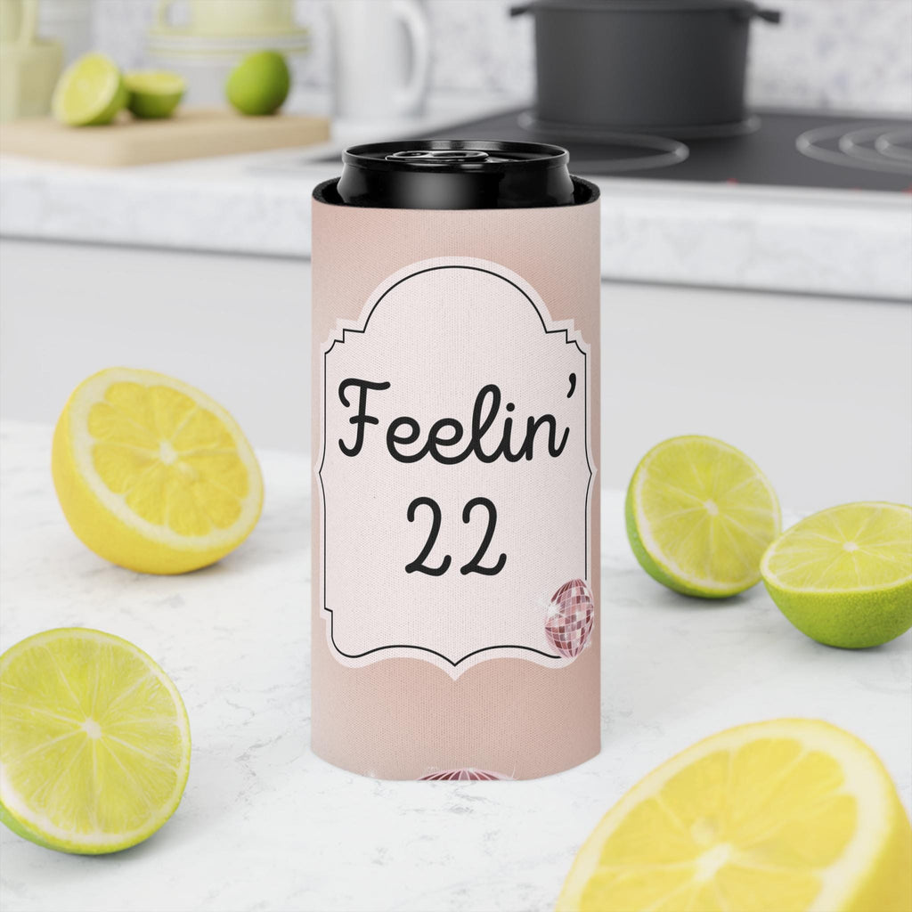 A pink can cooler sleeve with a black lid, featuring the text 'Feelin' 22' on a white label, set on a kitchen counter surrounded by fresh lemons and limes.