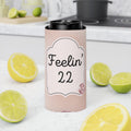 A pink can cooler sleeve with a black lid, featuring the text 'Feelin' 22' on a white label, set on a kitchen counter surrounded by fresh lemons and limes.