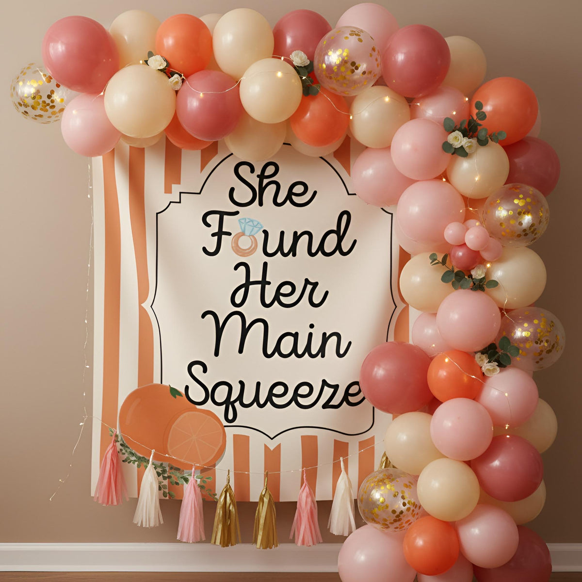 A decorative party backdrop with a balloon garland arch in blush pink, peach, cream, and metallic gold confetti balloons, surrounding a white sign that reads “She Found Her Main Squeeze” with a ring illustration. The setup is mounted against a beige wall with tassel accents at the bottom.