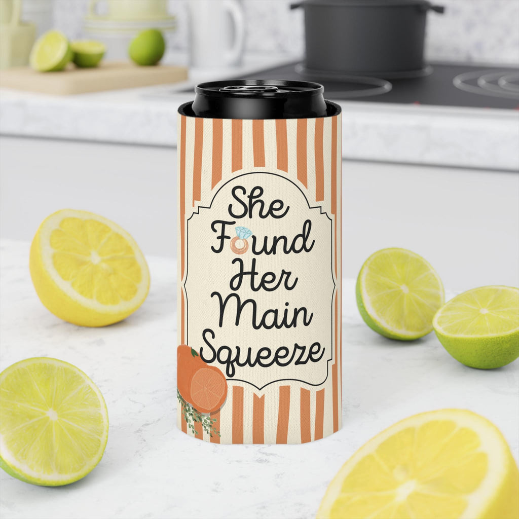 A slim can cooler sleeve with orange and white vertical stripes and a black lid, featuring the text 'She Found Her Main Squeeze' with a small ring graphic. The sleeve is shown on a can, styled on a white marble countertop with lemons and limes in a blurred kitchen background.