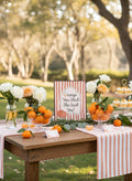 Outdoor wedding reception table setup with pink and white striped table runners, bowls of oranges, white and peach roses in glass vases, and a central sign reading 'Orange You Glad She Said Yes?' against a blurred park background.