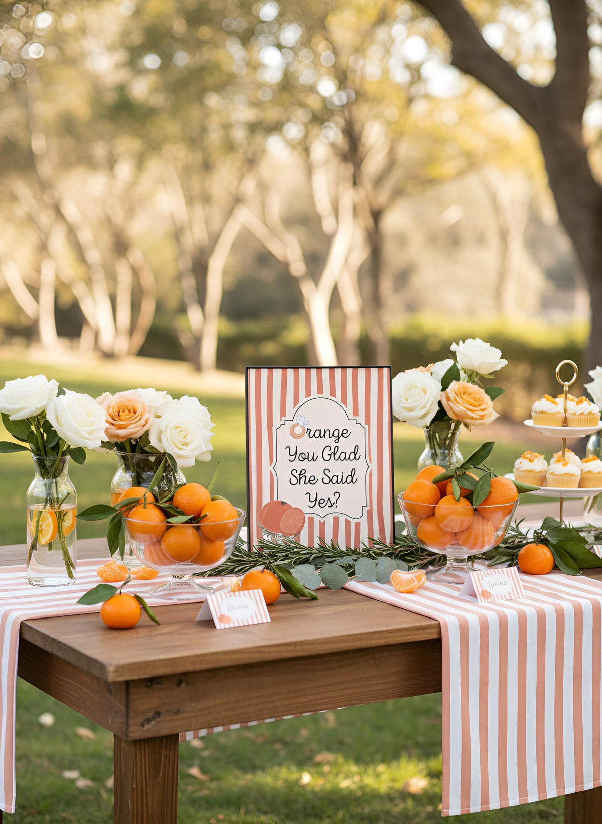 Outdoor wedding reception table setup with pink and white striped table runners, bowls of oranges, white and peach roses in glass vases, and a central sign reading 'Orange You Glad She Said Yes?' against a blurred park background.