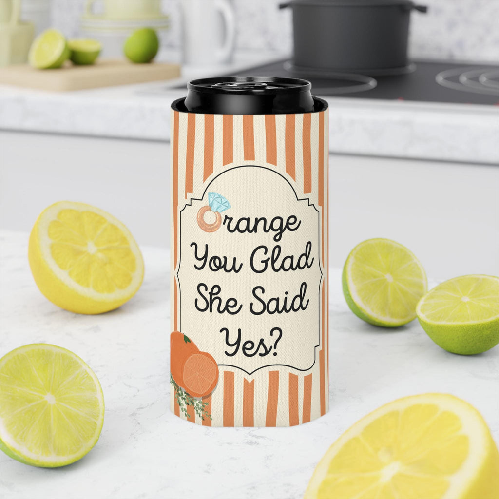 A slim insulated can cooler with a black lid, featuring orange and cream vertical stripes and a decorative label that reads 'Orange You Glad She Said Yes?' with an orange illustration. The cooler is styled on a white marble countertop with fresh orange slices and limes in a blurred kitchen background.