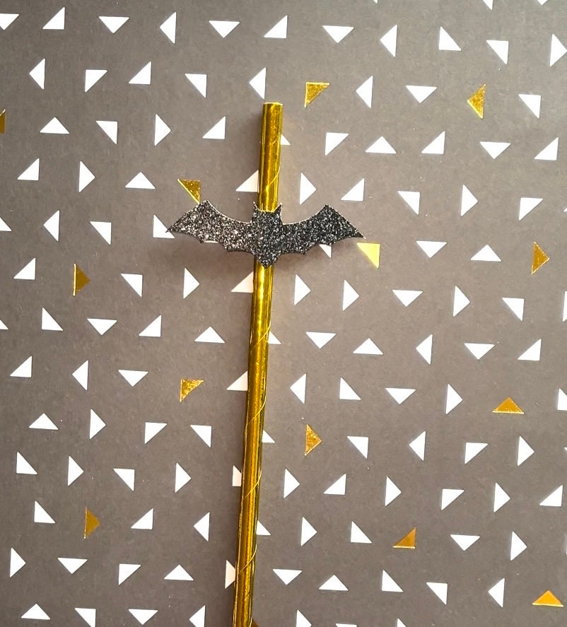 A Halloween-themed straw with a glittery black bat topper and a metallic gold tube, set against a gray patterned background with white and gold triangles.