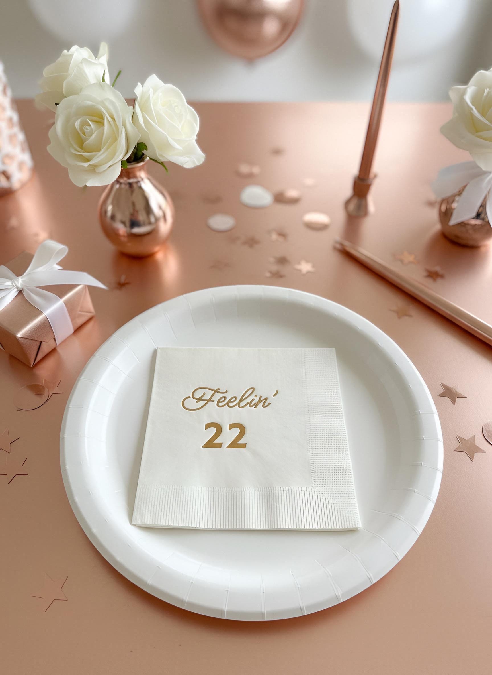 A styled party table setting featuring a white paper plate with a white napkin printed with 'Feelin' 22' in rose gold, silver, black, gold foil, surrounded by rose gold and white decorative elements including a candle, roses, and a gift box.