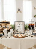 A styled baby shower or bachelorette party coffee station with a central sign reading 'A Baby Is Brewing,' white mugs, coffee bags, cookies on a wooden board, a glass jar of iced coffee, and small chalkboard signs on a cream tablecloth.