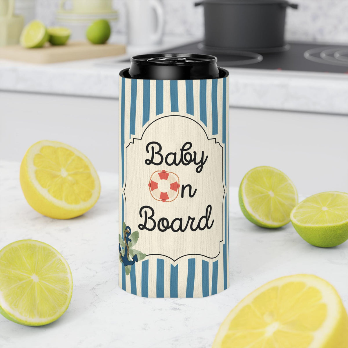 A tall insulated can cooler with a navy and white striped pattern and a cream label reading 'Baby on Board' with a small red-and-white life buoy icon. The cooler is shown on a kitchen counter surrounded by lemon halves and limes, with a stove and mug blurred in the background.