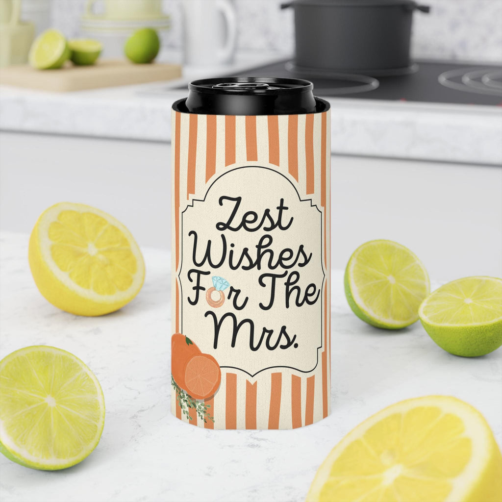 A slim can cooler sleeve with orange and white vertical stripes and the text 'Zest Wishes For The Mrs.' stands on a white countertop, surrounded by sliced lemons and limes, with a blurred kitchen background.