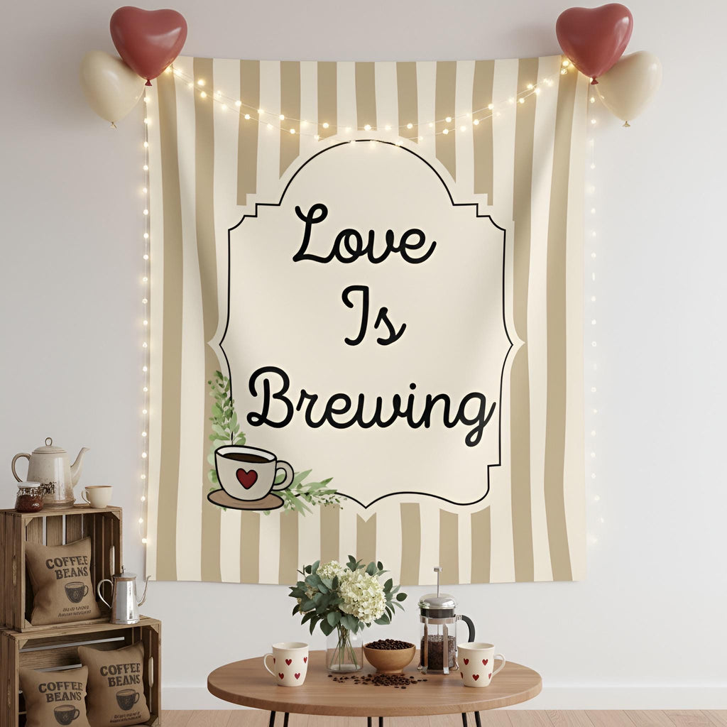 A decorative fabric wall tapestry hanging on a white wall, featuring the phrase 'Love Is Brewing' in black script inside an ornate cream label. The tapestry has beige and white vertical stripes and includes a small coffee cup illustration with a red heart.