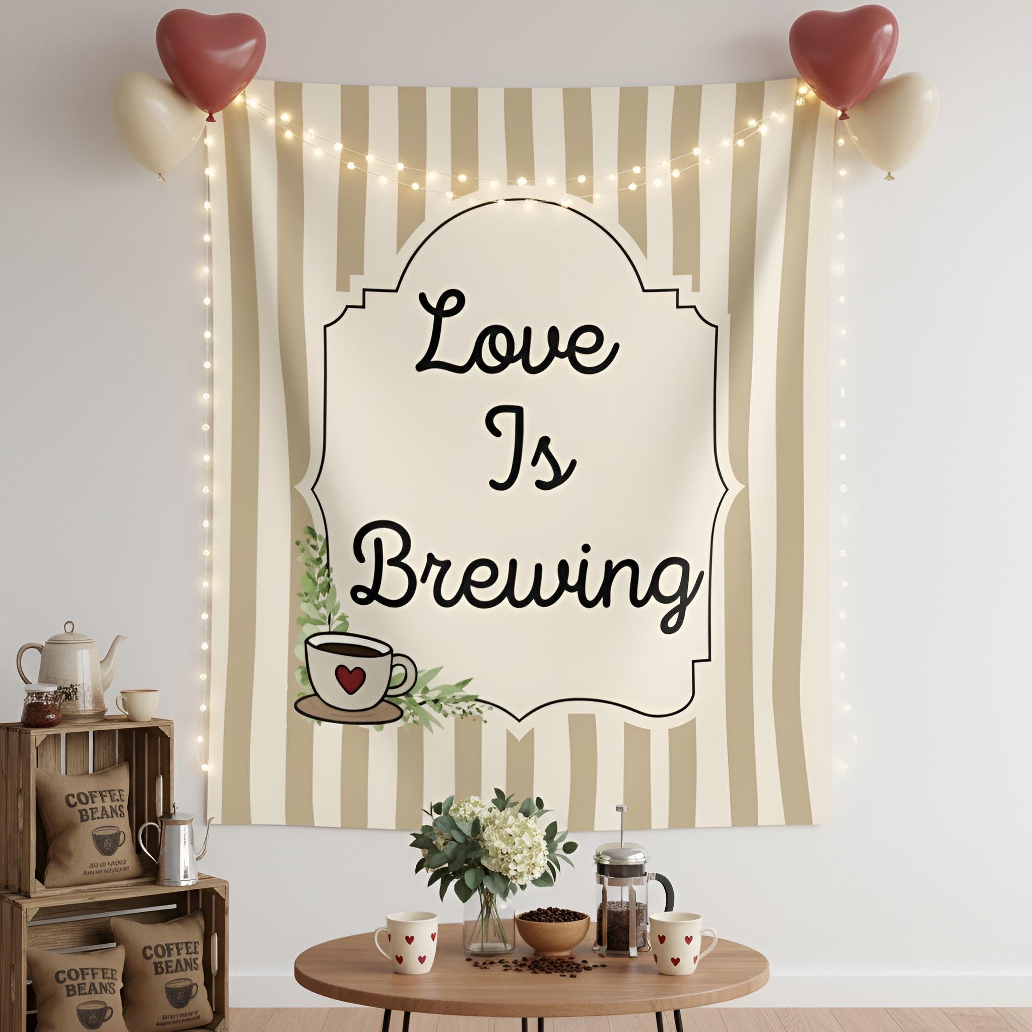 A decorative fabric wall tapestry hanging on a white wall, featuring the phrase 'Love Is Brewing' in black script inside an ornate cream label. The tapestry has beige and white vertical stripes and includes a small coffee cup illustration with a red heart.