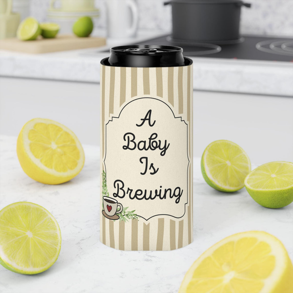 A stainless steel can cooler sleeve with a beige and white vertical stripe pattern and a central cream label that reads 'A Baby Is Brewing' with a small coffee cup illustration. The sleeve is shown on a black can, set on a white marble countertop surrounded by fresh lemon slices and limes, with a blurred kitchen background.