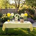 A styled outdoor party table setup in a garden with a pale yellow tablecloth, yellow and green balloons, small food stations, and our little lemon drop  branding