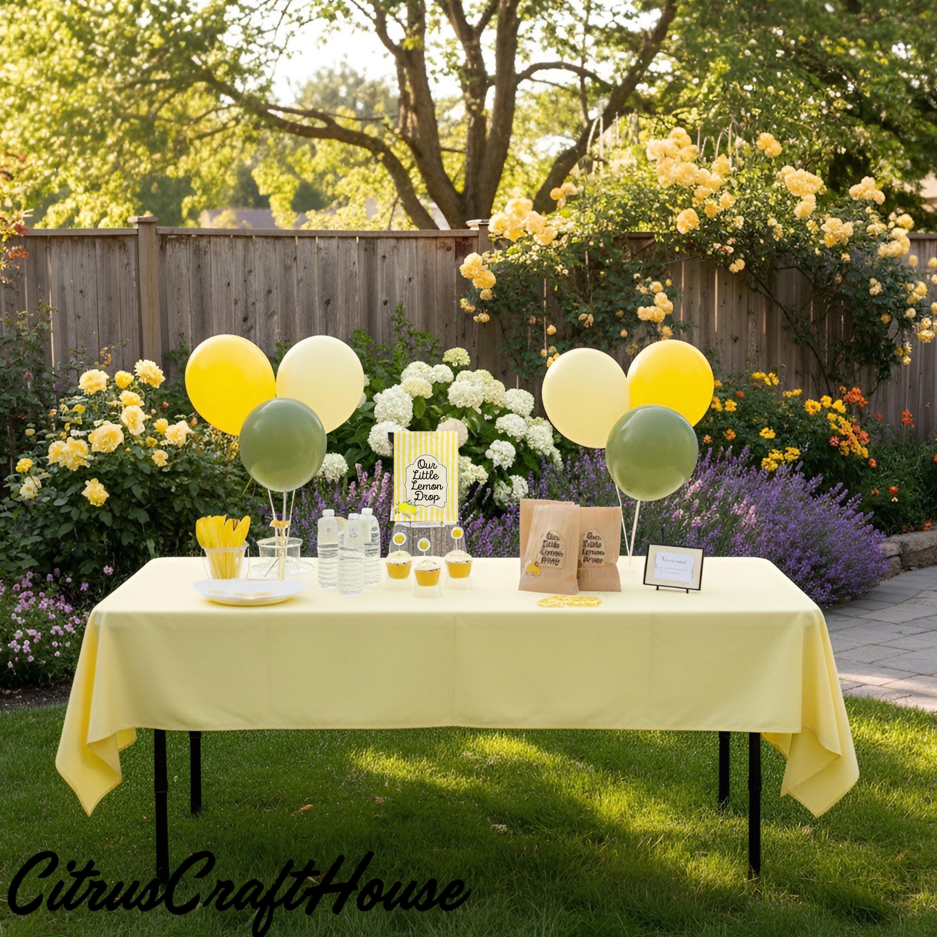 A styled outdoor party table setup in a garden with a pale yellow tablecloth, yellow and green balloons, small food stations, and our little lemon drop  branding