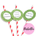 Three green glitter holiday straw picks with white circular inserts reading 'Merry Christmas,' 'STINK STANK STUNK!,' and 'You're a Mean One!' on red-and-white striped straws, shown on a white background with a pink 'Bestseller' badge.