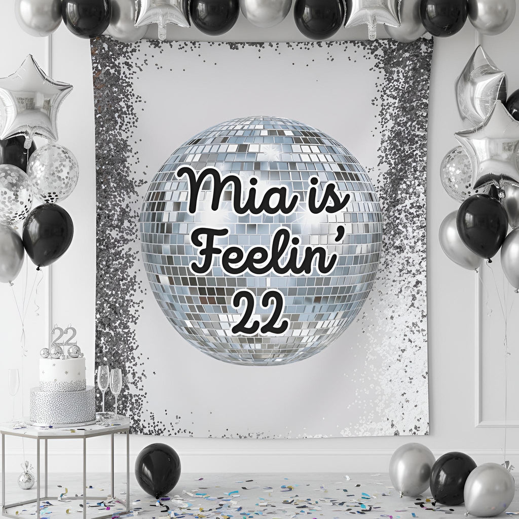 A silver and black party backdrop banner featuring a large disco ball graphic with the text 'Feelin' 22' in black script. The banner is styled in a party room setting with black and silver balloons, a silver and white tiered cake on a small table, and scattered confetti on the floor.