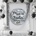 A silver and black party backdrop banner featuring a large disco ball graphic with the text 'Feelin' 22' in black script. The banner is styled in a party room setting with black and silver balloons, a silver and white tiered cake on a small table, and scattered confetti on the floor.