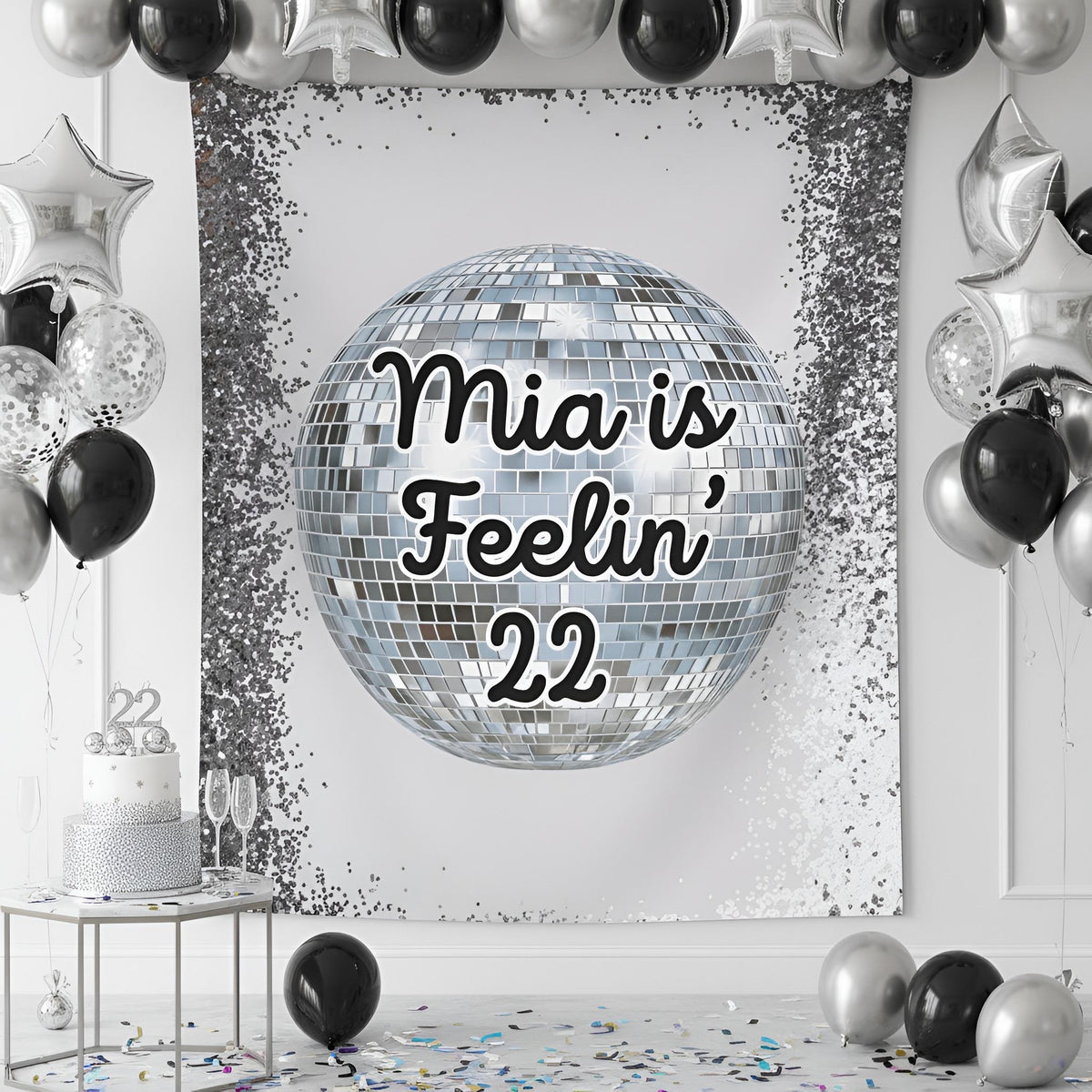 A silver and black party backdrop banner featuring a large disco ball graphic with the text 'Feelin' 22' in black script. The banner is styled in a party room setting with black and silver balloons, a silver and white tiered cake on a small table, and scattered confetti on the floor.