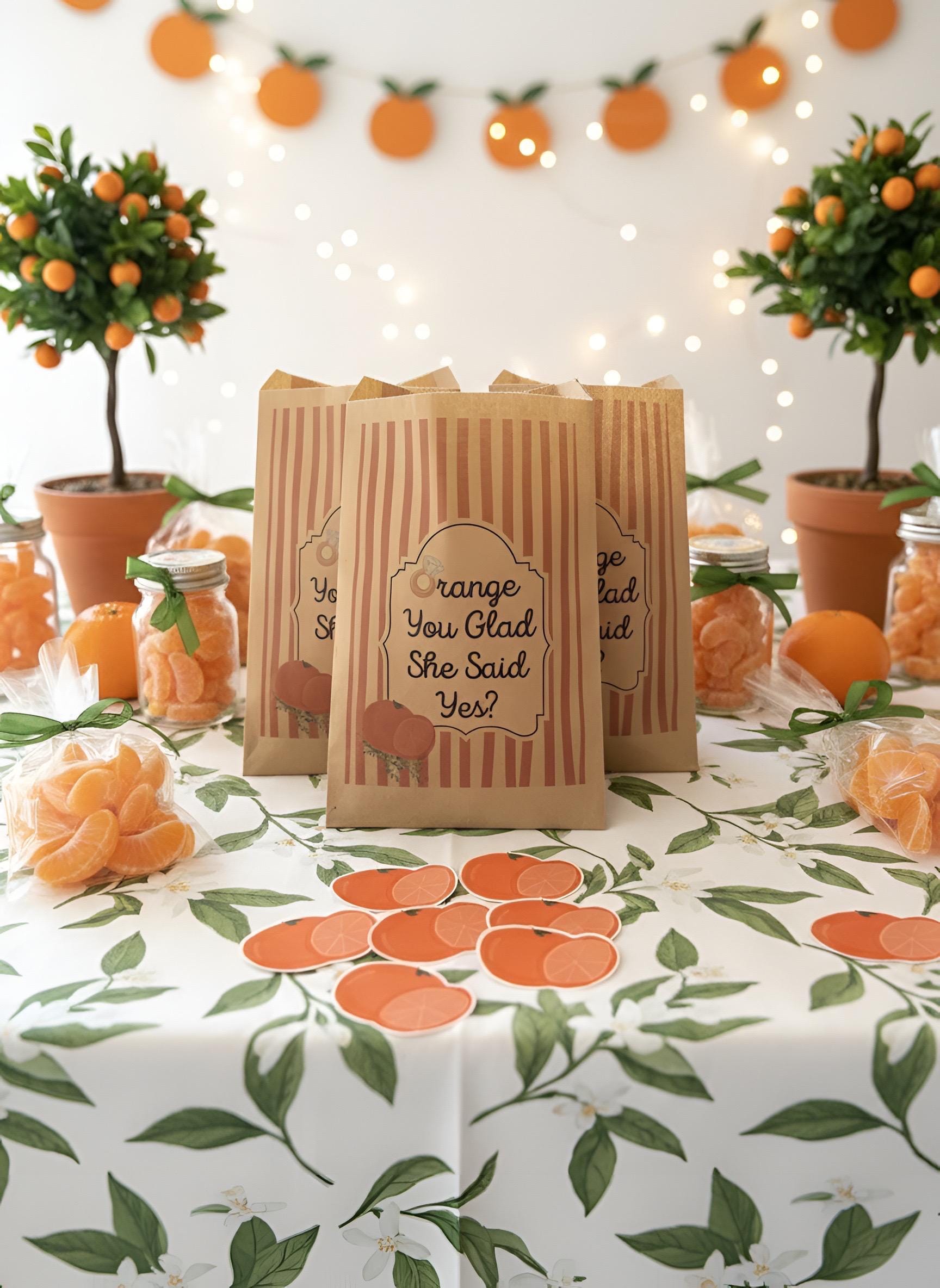 An orange-themed party table setup with three brown paper gift bags printed with orange slices and the phrase 'Orange You Glad She Said Yes?' in the center, surrounded by tangerines, jars, potted orange trees, and an orange leaf patterned tablecloth.