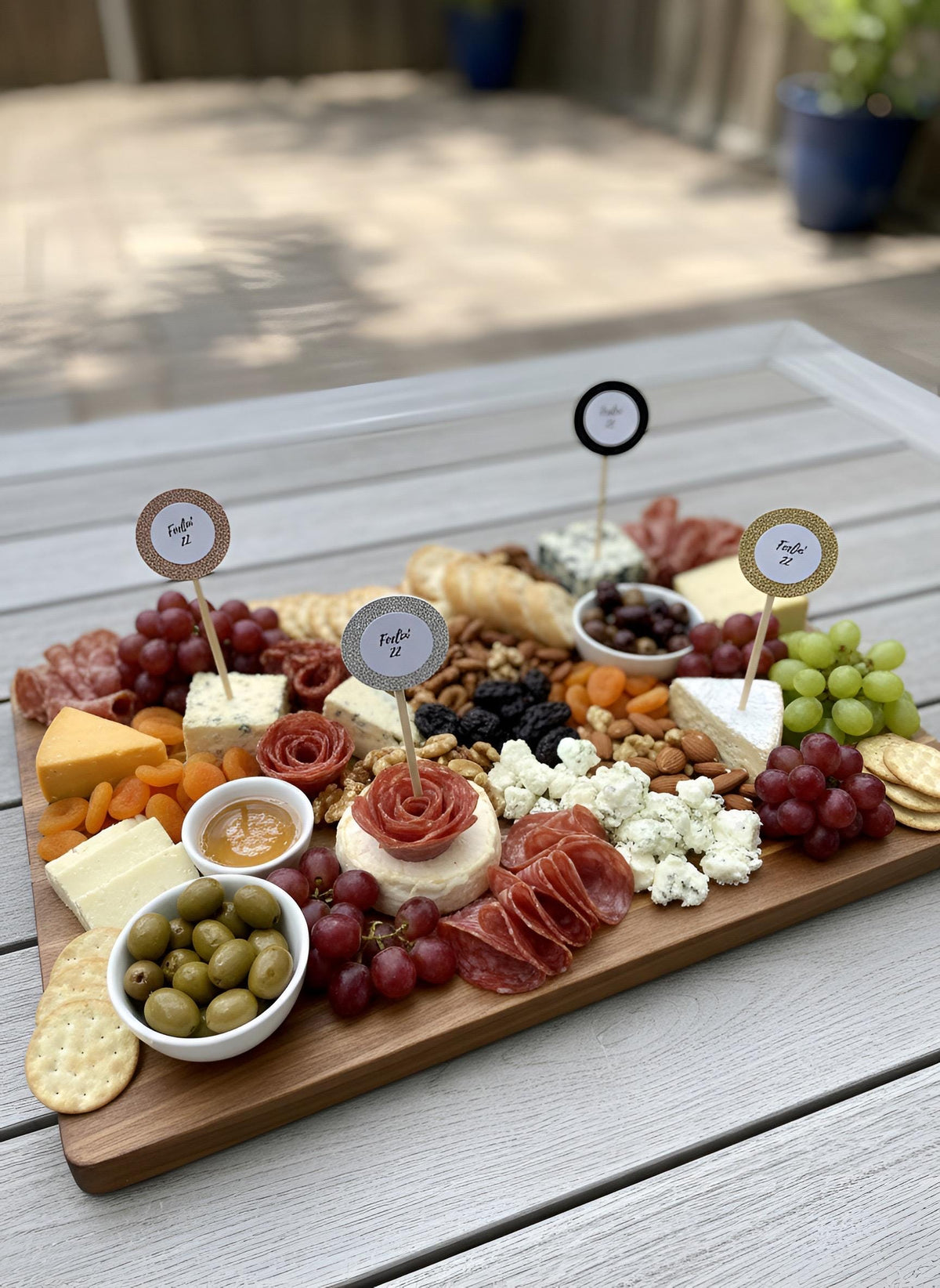 A wooden charcuterie board arranged outdoors on a light gray table, featuring cheeses, cured meats, grapes, olives, crackers, nuts, and small bowls of spreads. Rose gold, gold, silver, black glitter, treat, toppers, cupcake pics, charcuterie, pics with feeling 22 written on it