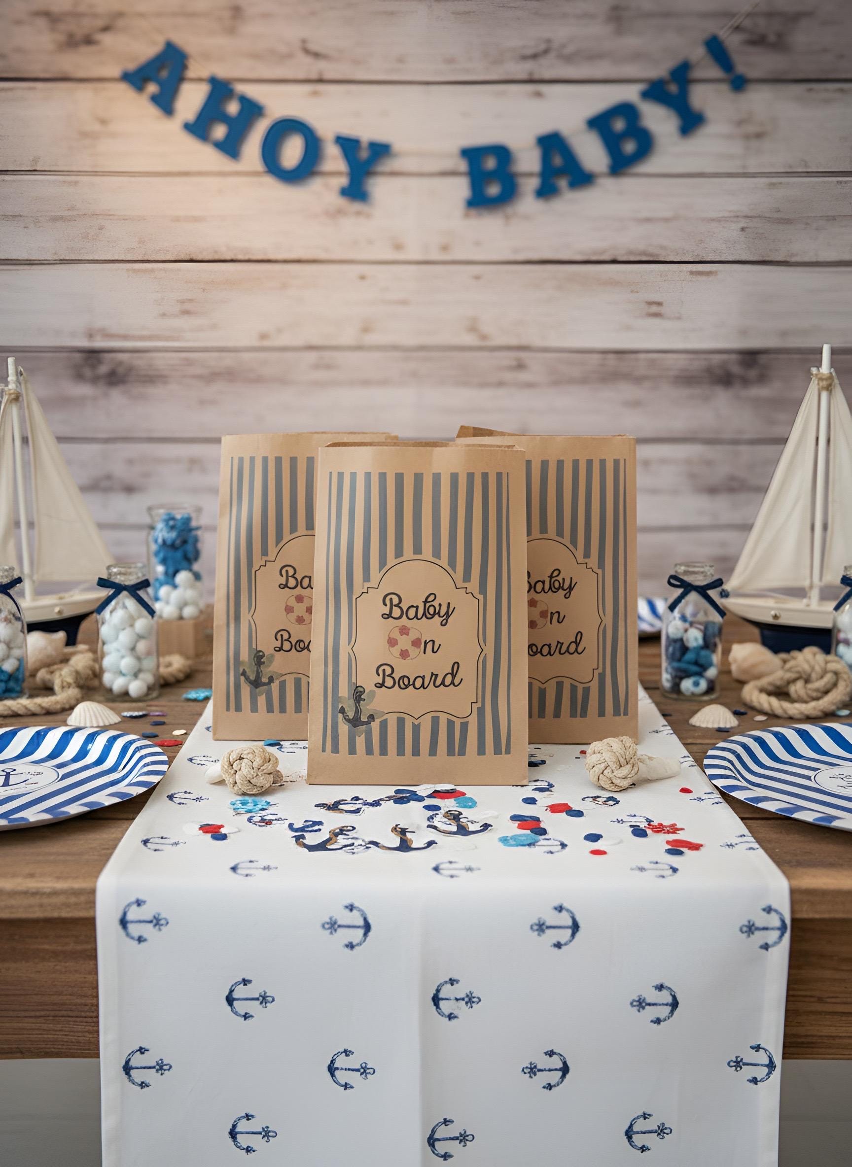 Nautical-themed baby shower party supplies displayed on a wooden table, including kraft paper bags with blue stripes and 'Baby on Board' labels, a white table runner with blue anchors, striped plates, and wooden model sailboats against a wood plank wall with a blue 'AHoy BABY!' banner.