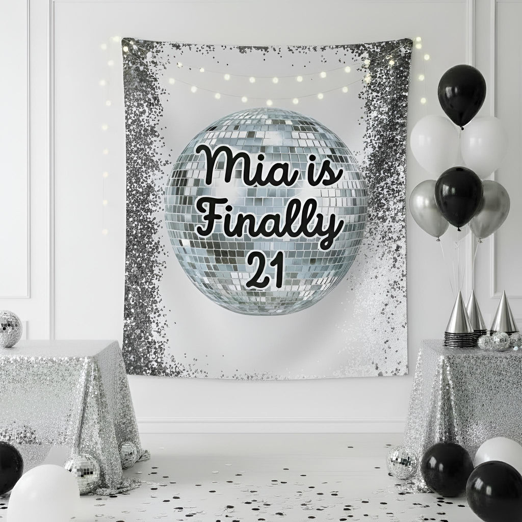 A silver and white party tapestry featuring a disco ball graphic with the text ‘Finally 21' in black script, displayed on a white wall in a decorated room with black and white balloons, silver tablecloths, and confetti.