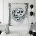 A silver and white party tapestry featuring a disco ball graphic with the text ‘Finally 21' in black script, displayed on a white wall in a decorated room with black and white balloons, silver tablecloths, and confetti.