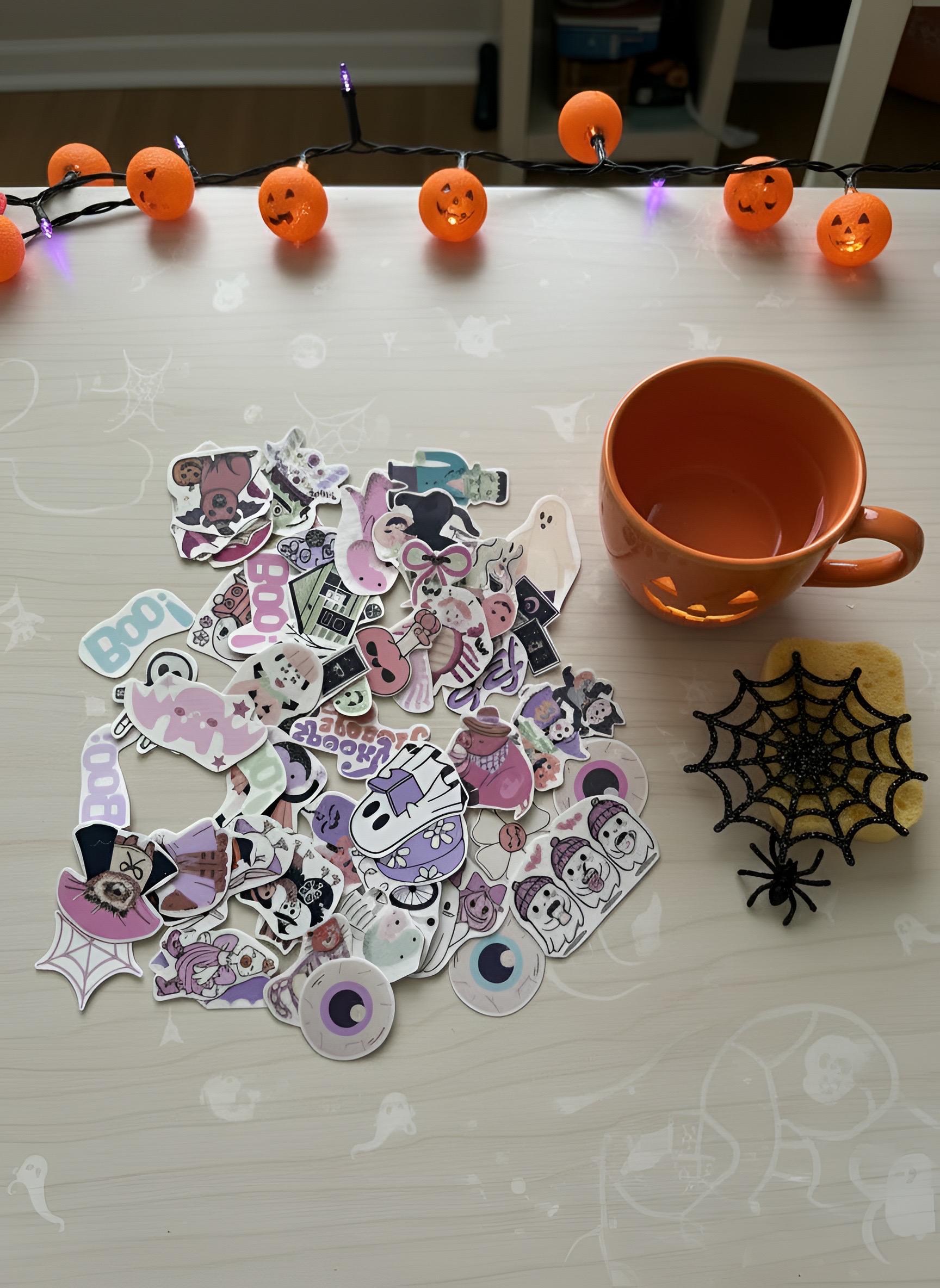 A pile of Halloween-themed sticker temporary tattoo cutouts in pastel colors, arranged on a tabletop next to an orange mug, a black spiderweb decoration, and orange pumpkin string lights.