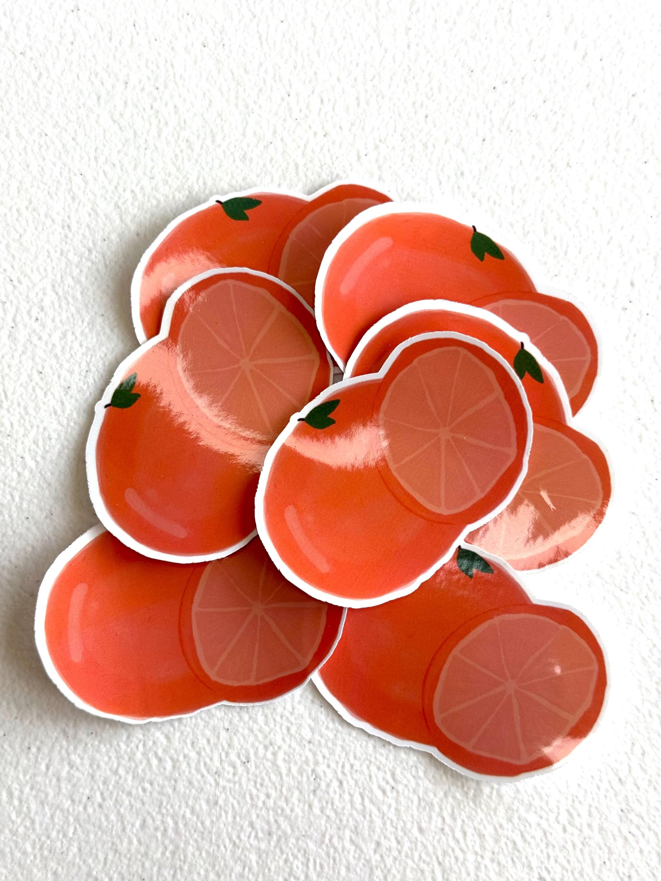 A pile of die-cut stickers shaped like sliced oranges, featuring a glossy orange-red surface with a white border, set against a textured off-white background.