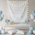 A styled mahjong setup featuring a large white wall banner with light blue decorative borders, champagne glass illustrations, and the text 'Mahjong & Mimosas,' surrounded by warm string lights. A white table below holds a mahjong deck, two glasses with amber drinks, and flower arrangements, while blue and white balloons decorate the scene.