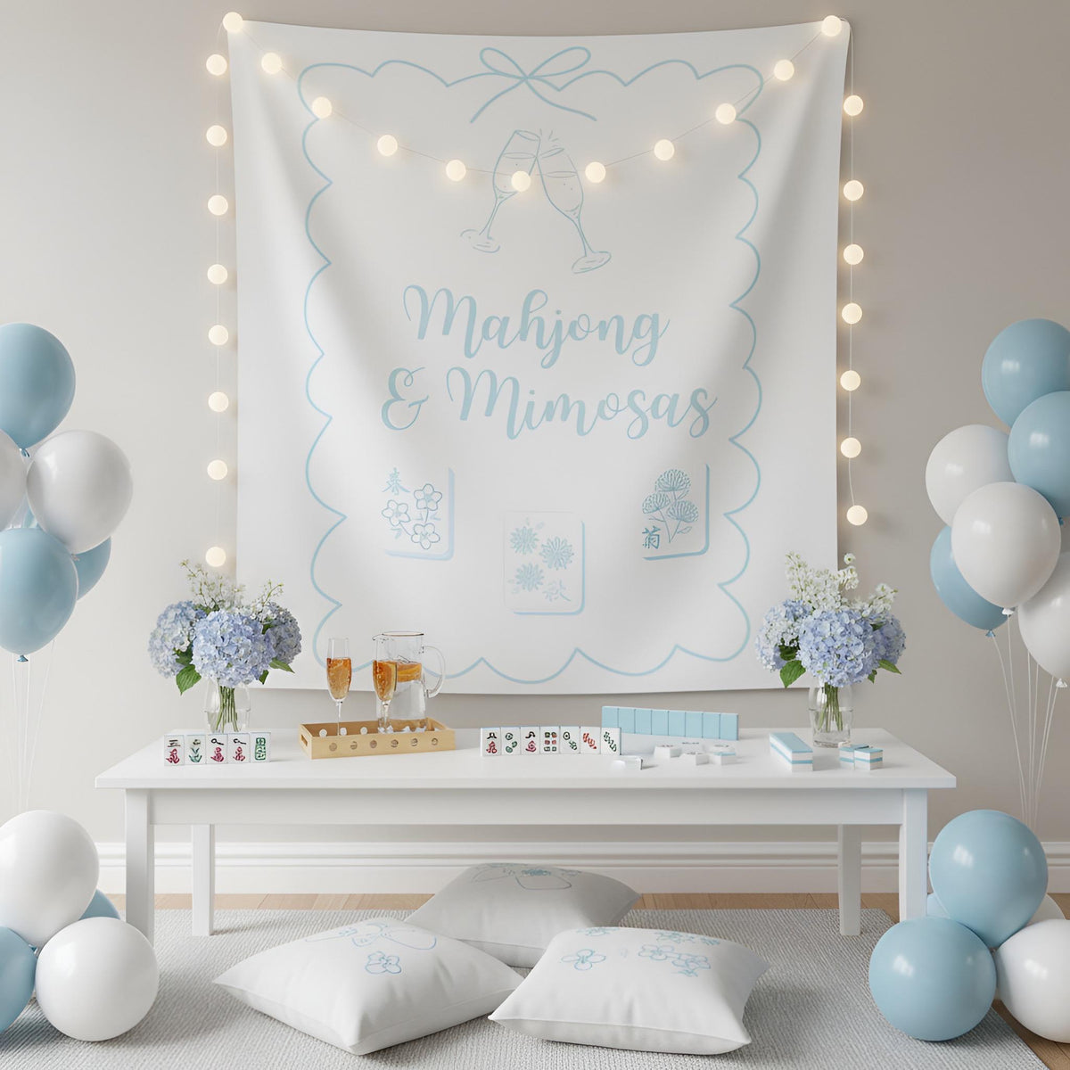 A styled mahjong setup featuring a large white wall banner with light blue decorative borders, champagne glass illustrations, and the text 'Mahjong & Mimosas,' surrounded by warm string lights. A white table below holds a mahjong deck, two glasses with amber drinks, and flower arrangements, while blue and white balloons decorate the scene.
