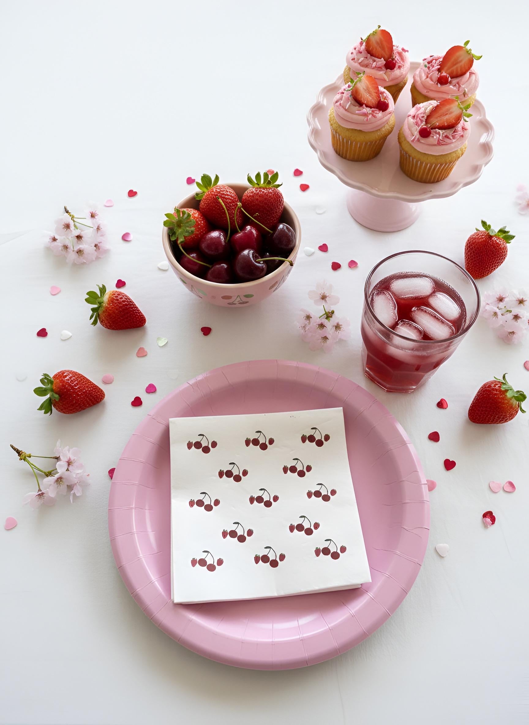 A styled tabletop scene with pink paper plates, a napkin printed with cherries, strawberries, a glass of pink iced drink, cupcakes on a pink stand, and a bowl of cherries on a white background with scattered heart confetti and flowers.