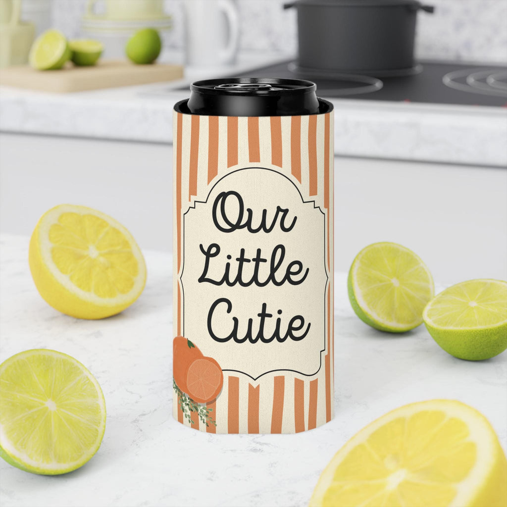A tall insulated can cooler sleeve with orange and cream vertical stripes and a black lid, printed with the phrase 'Our Little Cutie' and a small orange illustration. The sleeve is shown in a kitchen setting with lemons and limes around it.