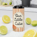 A tall insulated can cooler sleeve with orange and cream vertical stripes and a black lid, printed with the phrase 'Our Little Cutie' and a small orange illustration. The sleeve is shown in a kitchen setting with lemons and limes around it.