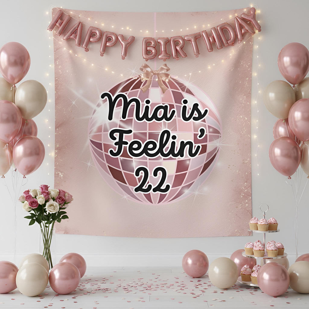 A pink and rose gold birthday party backdrop banner featuring a disco ball graphic with the text 'Feelin' 22' in black script. The banner is decorated with a 'HAPPY BIRTHDAY' balloon garland, surrounded by pink and cream balloons, cupcakes on a tiered stand, and a vase of roses.