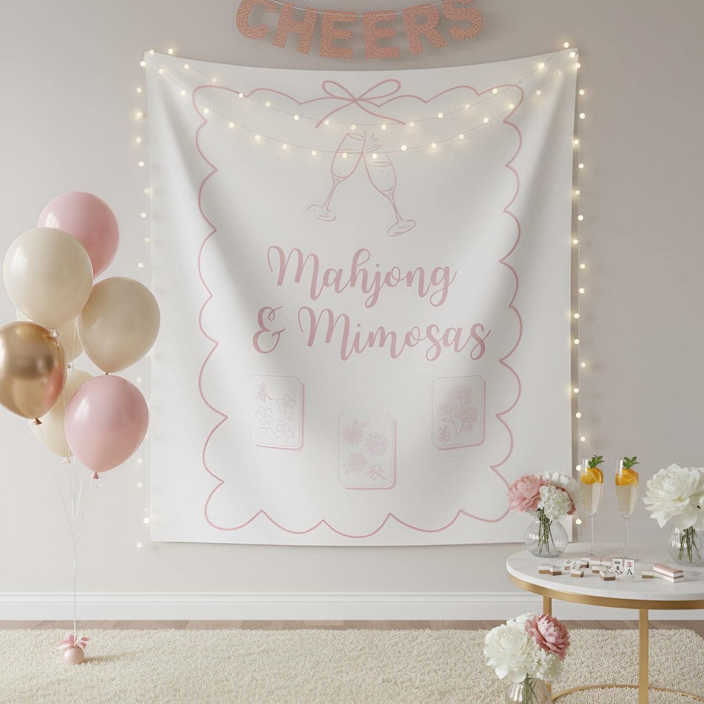 A white fabric party backdrop banner with pink line art and text reading 'Mahjong & Mimosas,' hung on a wall in a decorated room with string lights, balloons, flowers, and a small table.
