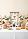 A styled coffee bar setup with two cups of latte, small bowls of coffee beans, cupcakes on stands, flowers, and a central sign that reads 'Love Is Brewing' on a white table against a warm beige background.