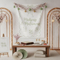 A styled wedding or event booth setup featuring a large white fabric banner with 'Mahjong & Matrimony' in sage green script, decorated with cherry blossom garlands, hanging ornaments, and string lights. A wooden table below holds game pieces, snacks, and two champagne flutes, with folding fans and cushions on the floor.