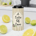 A tall insulated tumbler with a black lid stands on a white countertop in a kitchen setting. The tumbler sleeve has beige and cream vertical stripes and a central label that reads “A Latte Of Love” with a small coffee cup illustration.