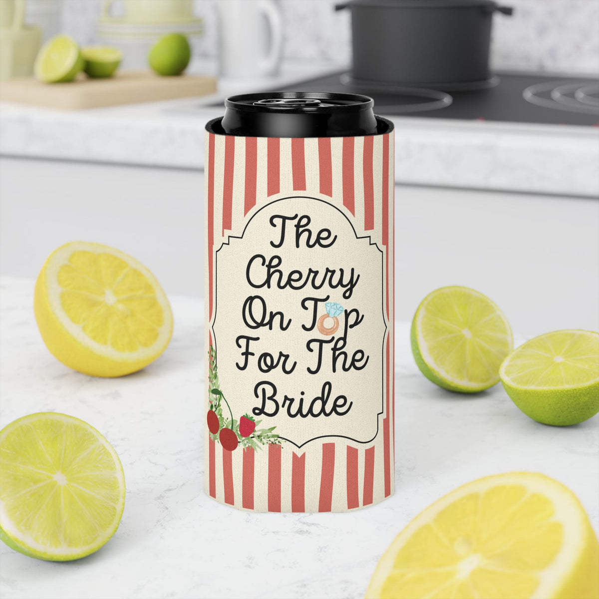 A slim can cooler sleeve with red and white vertical stripes and a cream label reading 'The Cherry On Top For The Bride,' shown around a black can on a kitchen countertop with lime slices around it.