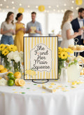 A yellow and white striped sign with the phrase 'She Found Her Main Squeeze' in black script, displayed on a decorated party table with lemons, flowers, and blurred guests in the background.